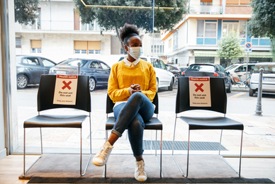 Teen wearing mask sitting alone flanked by 2 empty chairs marked for social distancing 